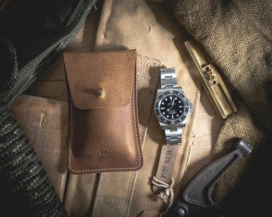 A silver wristwatch with a black dial rests on a beige cloth beside the Rugged Chestnut brown leather pouch, displaying beautifully patinaed leather, a brass whistle, and a folded fabric. The initials "BO" are engraved on both the pouch and the whistle, adding a touch of personalization to this rustic, outdoorsy scene.