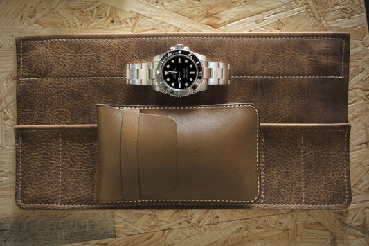 A silver wristwatch with a black dial is placed on an unfolded brown watch roll made from Old-Olive veg tan leather. Resting on a textured wooden surface, it's accompanied by a small Sample Sale pouch, showcasing the elegance of personalized accessories.