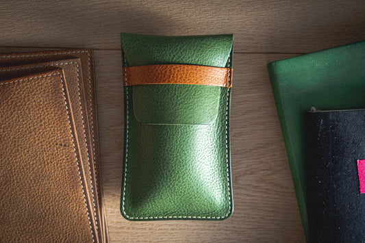 A SAMPLE SALE Pebbled veg-tan eyeglass case in lime with a cognac strap is placed on a light wooden surface. It is surrounded by two brown leather pieces on the left and a green-and-black book on the right. The eyeglass case is positioned vertically in the center of the image.