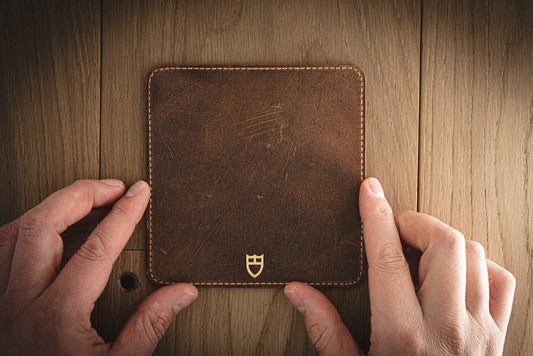 Two hands hold the SAMPLE SALE Valet, a vintage brown leather coaster measuring 13 x 13 cm. It rests on a wooden surface, featuring a small embossed emblem near the bottom edge. The coaster has a slightly weathered appearance with visible creases that add character, reminiscent of exclusive sample sale items.