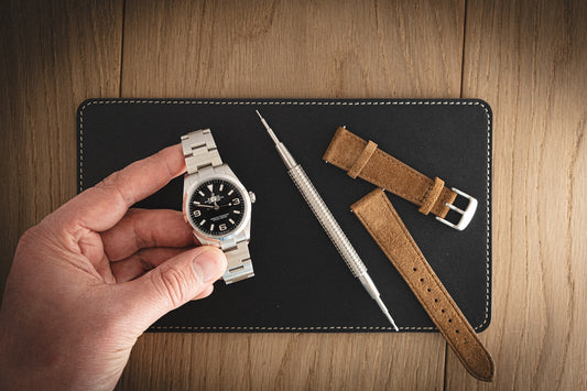 A hand displays a silver watch above a dark blue leather SAMPLE SALE Valet (13 x 23 cm) on a wooden surface. Nearby, there's an elegant arrangement of a watch strap replacement tool along with a brown leather watch strap.