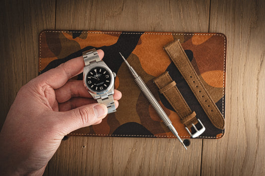 A hand holds a silver wristwatch with a black face over an orange (dark) camo leather Valet measuring 13 x 23 cm, suggesting its robust style. Nearby, a brown leather watch strap and metal adjustment tool rest on the wooden background, complemented by the sophistication of suede. Perfect for discovering in a SAMPLE SALE.