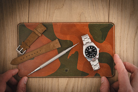 An orange camouflage-patterned leather valet from the SAMPLE SALE collection is held in hand, featuring a stainless steel wristwatch with a black face, an interchangeable brown leather strap, and a metal watch tool. The wooden surface beneath complements the rustic aesthetics.