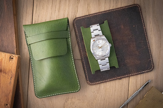 A silver wristwatch with a metal band is displayed on green fabric atop a brown leather square, next to a closed SAMPLE SALE green, full leather case protector. The elegant setting features a light wooden surface and various wooden materials.