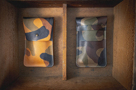 Two pouches from the Veg-tan CAMO COLLECTION with camouflage patterns are displayed side by side on a wooden shelf. The pouch on the left, crafted from veg-tan leather, showcases a mix of tan, black, and brown shades. Meanwhile, the pouch on the right features green, brown, and black hues. Each handmade watch pouch includes a flap closure.