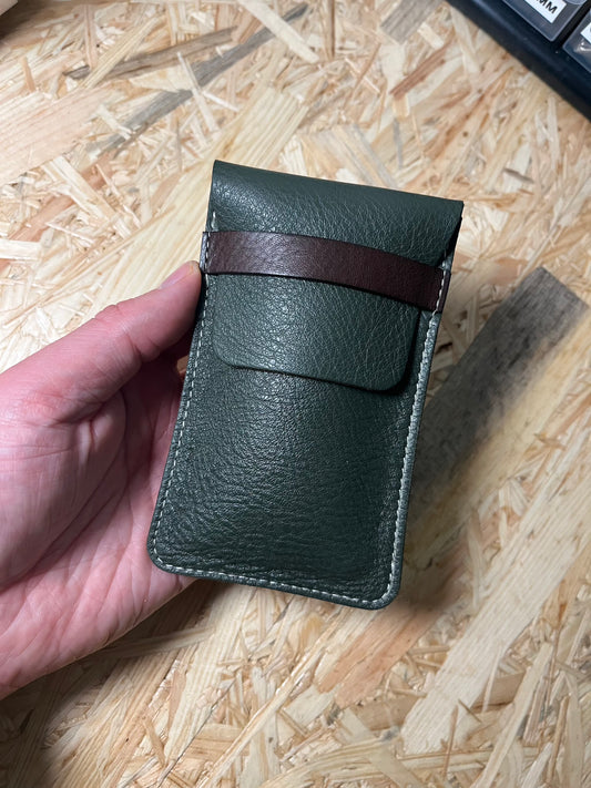 A hand showcases the SAMPLE SALE dark green phone pouch with a brown strap and white stitching from the Secret Sale Department. This exclusive item rests against a wooden background, offering a glimpse into the world of sample-made pouches.