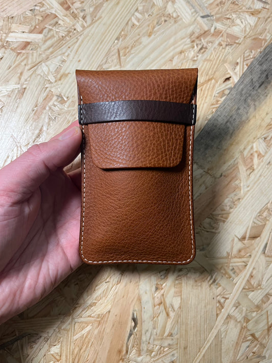 A hand holds a SAMPLE SALE pouch made from veg tan leather in cognac, featuring a dark brown strap and fold-over flap. The background displays a textured light wood surface, emphasizing the artisanal appeal of these sample-made pouches.