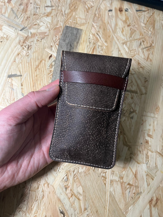 A hand holding the SAMPLE SALE coffee brown premium pouch, equipped with a brown strap, rests on a textured wooden surface.