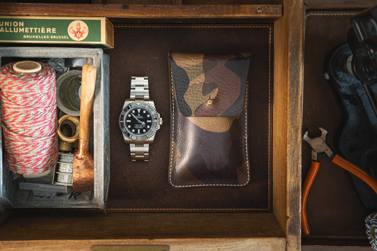 A drawer with neatly organized items including a spool of twine, a wooden-handled hammer, a watch with a metal band, pliers, and a small box labeled "Union Allumettière Bruxelles-Brussel," along with the SAMPLE SALE Green camo stiff veg tan leather STUD from the Secret Sale Department.