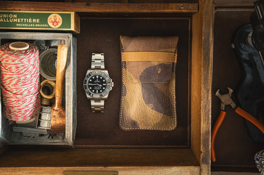 A wooden drawer organized with various items: a roll of red and white twine, a wooden-handled tool, a Swiss wristwatch with a metallic band, a SAMPLE SALE Green camo (golden WP logo small) leather pouch made from stiff veg tan leather, a pair of pliers, and a box labeled 'Union Allumettiere'.