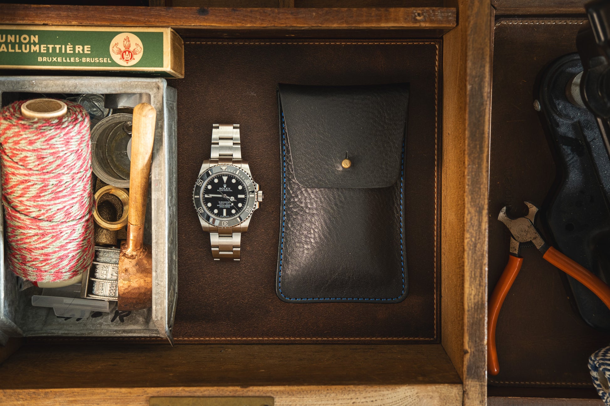 A top-down view of a wooden drawer reveals a wristwatch, a black pebbled leather pouch with soft veg tan leather and blue stitching, twine, a small mallet, and some tools. The items are neatly arranged with an organized appearance, highlighting the level of personalization involved in curating such details.