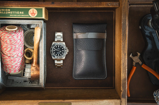 A vintage wooden drawer containing a coiled string, a brass hammer, a metal watch with a black face, an Earth tones Ocean blue pebbled veg-tan strap from the SAMPLE SALE collection, and orange-handled pliers. A green and yellow box labeled "Union Allumettière Bruxelles" is also visible.