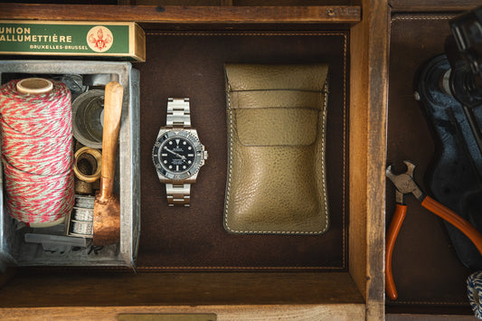 A wooden drawer contains various items, including a silver wristwatch, a SAMPLE SALE Pebbled veg-tan collection "Earth tones" Silver tusk leather pouch, a box of matches, a roll of twine, a wooden-handled magnifying glass, and pliers. The items are neatly arranged, showcasing a blend of vintage and modern tools.