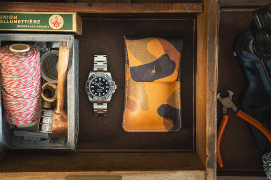 A wooden drawer contains various items: a neatly coiled red and white string, a pipe, tape, a stainless steel watch, an orange camo soft veg tan leather STUD from the SAMPLE SALE collection, a pack of matches, and a pair of orange-handled pliers.