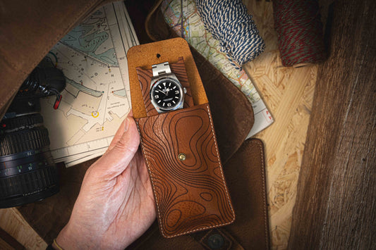 A hand holds an open Pathfinder case from the Pebbled veg-tan collection, exquisitely handmade in Antwerp, revealing a wristwatch with a black dial and silver bracelet. In the background, a camera lens, map, and spools of twine evoke a sense of travel or adventure.