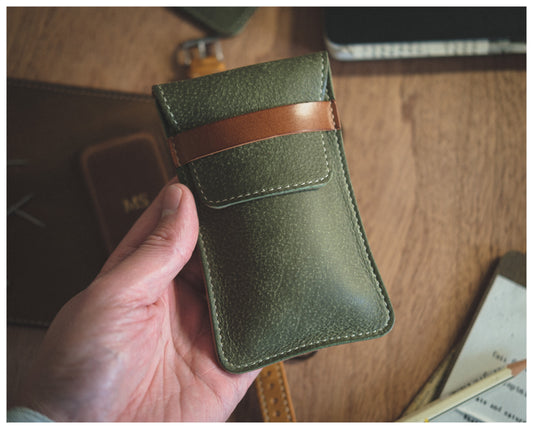 A hand holds a green Soft Layered Premium Pouch (with lining) featuring a brown strap across the top. Handmade in Antwerp, this high-end watch accessory rests on a wooden surface surrounded by other leather items, a notebook, and blurred papers in the background.