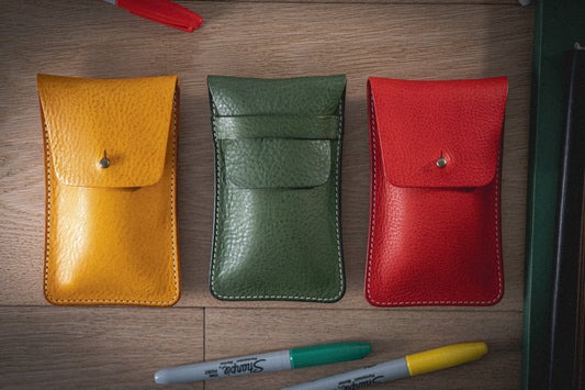 Three pouches from the Pebbled Veg-Tan Collection "Vibrant" in yellow, green, and red are displayed on a wooden surface. Handmade in Antwerp from pebbled veg-tan leather, each pouch features a snap closure. Below them, two Sharpie markers—one with a red cap and the other with a green cap—are partially visible. Additional pens and parts of a book frame the pouches.