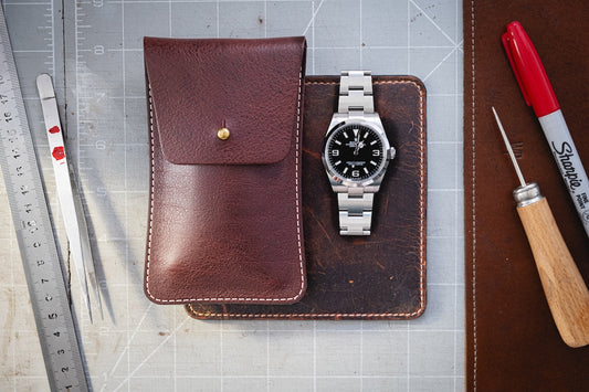 A flat lay image features a vintage Burgundy brown leather watch pouch, a silver wristwatch with a black dial, rulers, a red marker, and a leatherworking tool artfully arranged on a grid-patterned cutting mat.