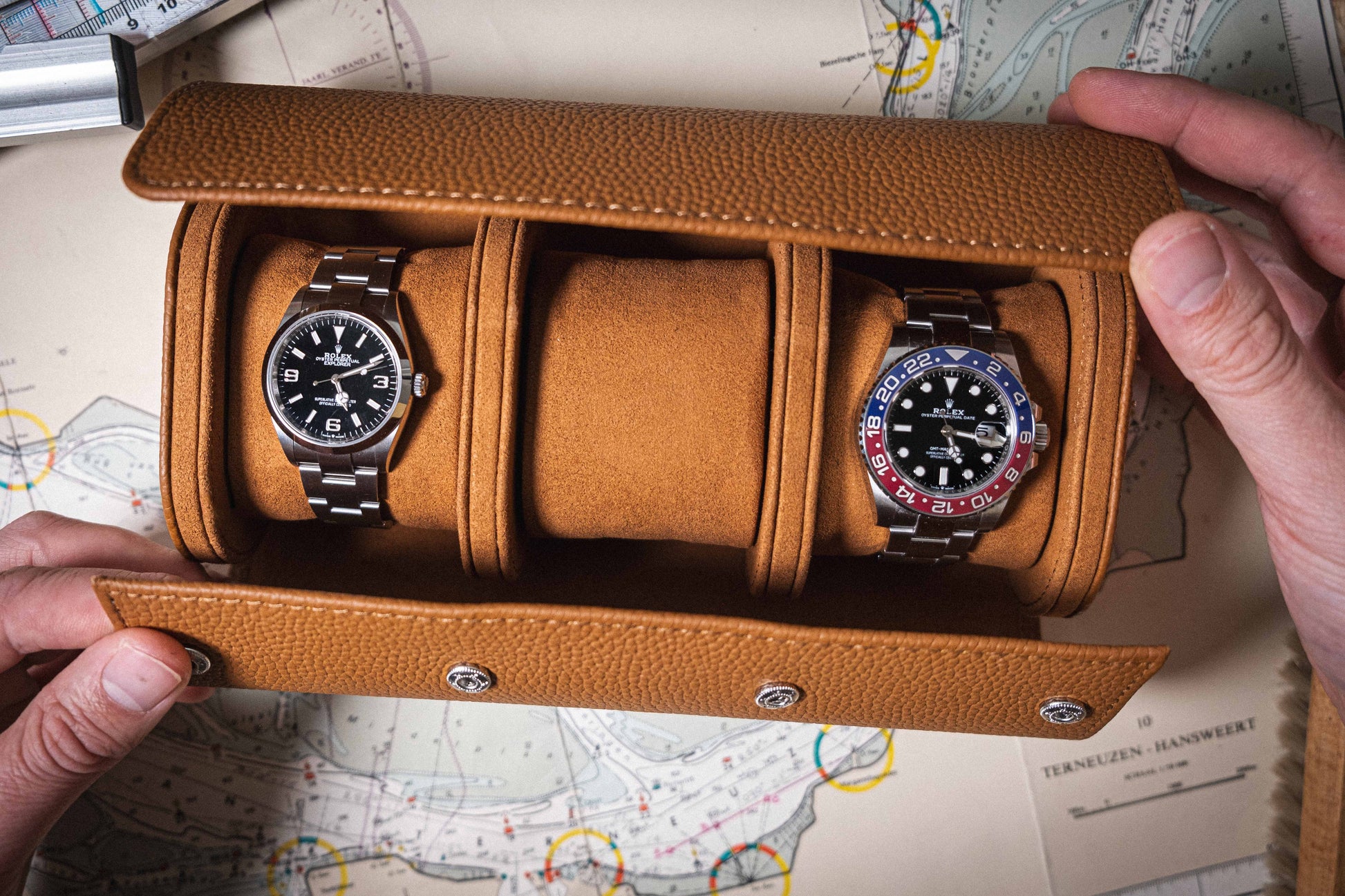 A person holds an open Oval Travel Case made of full-grain leather, displaying two wristwatches: one featuring a black face with a silver bezel and the other boasting a blue and red bezel. This sophisticated watch case is casually laid over a map in the background.