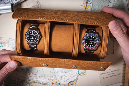 A person holds an open Oval Travel Case made of full-grain leather, displaying two wristwatches: one featuring a black face with a silver bezel and the other boasting a blue and red bezel. This sophisticated watch case is casually laid over a map in the background.