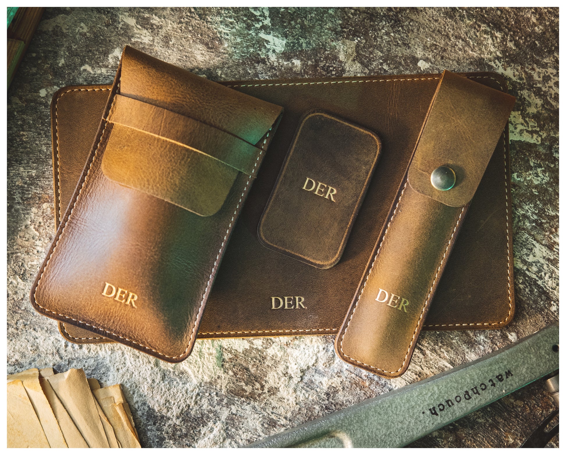 A collection of vintage brown leather accessories from the PACKAGEDEAL line, all embossed with the word "DER," is displayed on a textured surface. The collection includes a pouch, a small case, a long holder with a button closure, and a flat rectangular piece. A watch pouch distinctly marked with "waterpouch" stands out prominently at the bottom.