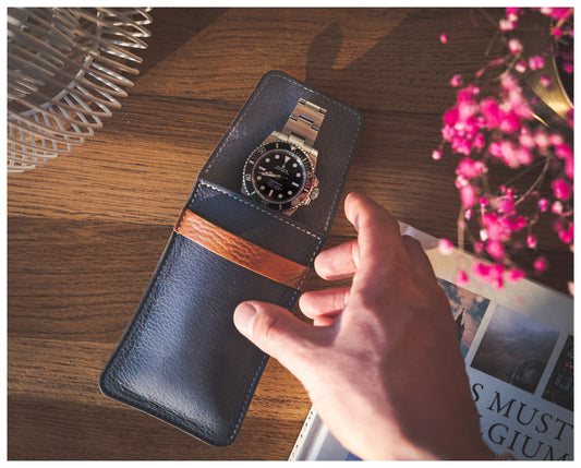 A hand reaches for a silver watch with a black dial inside an Ocean Blue soft layered premium pouch with lining, featuring a brown strap, all placed on a wooden table. Beside the pouch, there is a partially visible white magazine and a glass decorative item. Faintly in the background, pink flowers can be seen.