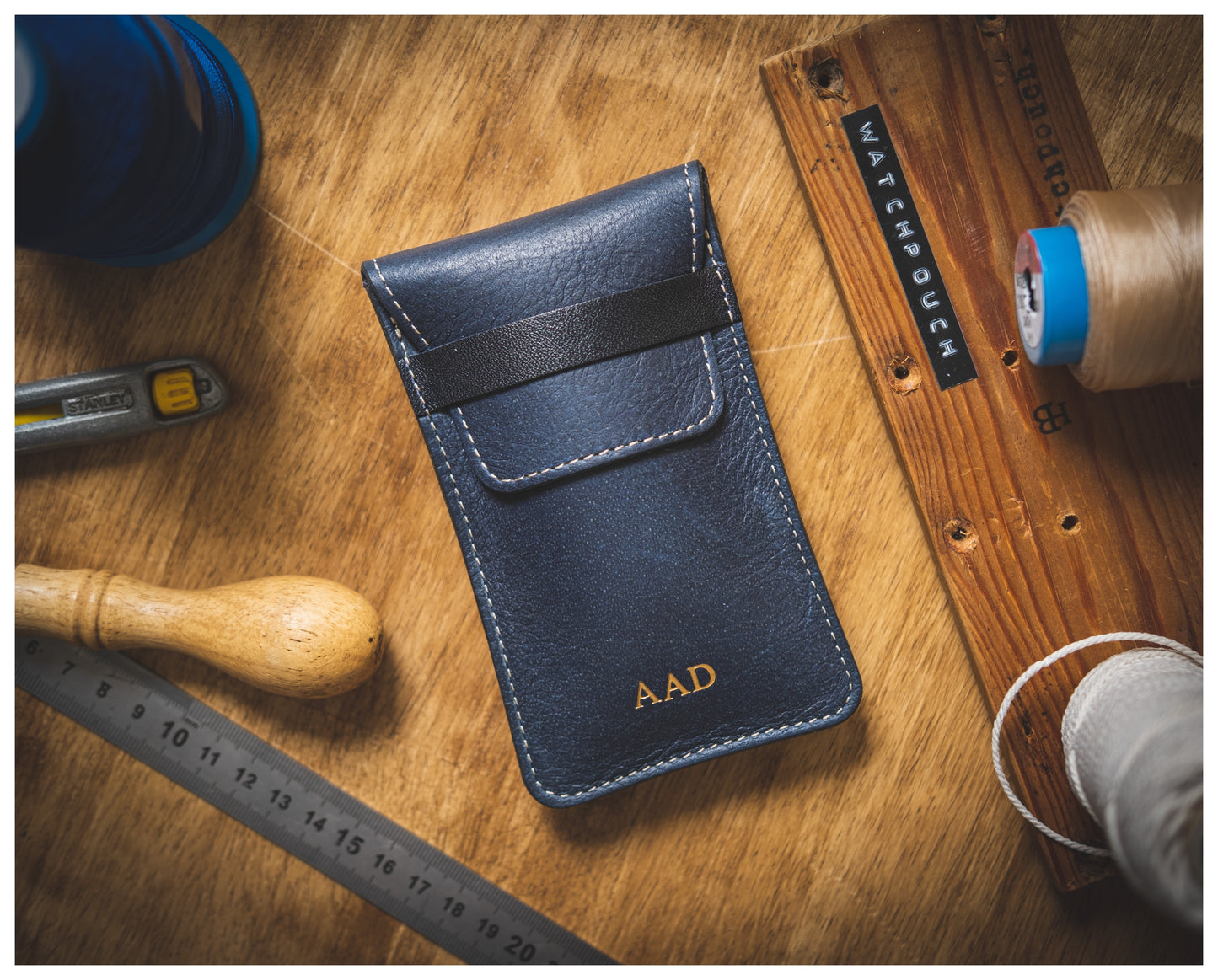 An Ocean blue (blue strap) soft layered premium pouch with a black strap and the initials "A.A.D" embossed in gold letters is placed on a wooden surface, surrounded by sewing tools such as thread spools, a wooden ruler, a tape measure, a tool handle, and a metal ruler. This handmade watch accessory hails from Antwerp.