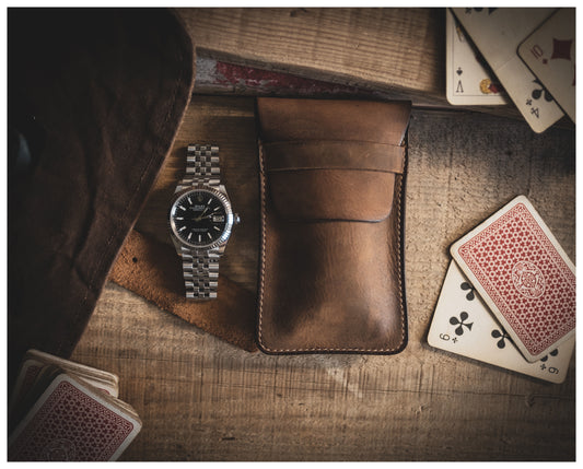 A stainless steel wristwatch with a black dial is placed next to the Product Name "vintage brown" watchpouch on a wooden surface. Playing cards, including an Ace of Diamonds and a Nine of Clubs, are scattered around the items.